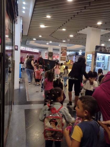 Crianças caminhando em fila por um corredor do shopping, acompanhadas pelas educadoras