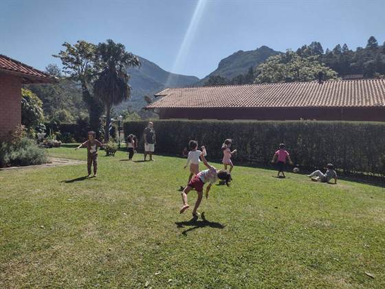 Crianças brincando no jardim, com casas de telhado colonial e montanhas ao fundo