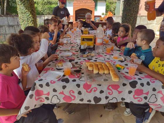 Crianças reunidas em volta de uma mesa comprida, lanchando pão de salsicha e suco sob uma área coberta