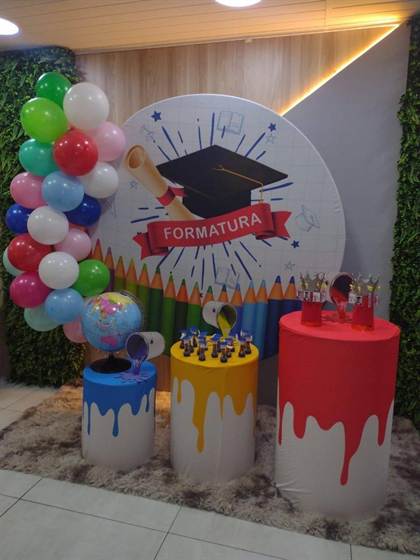 Painel de formatura com capelo, diploma e lápis de cor, e mesas com tema de tinta colorida