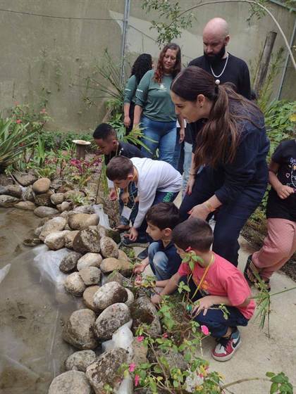 Crianças e adultos observam um lago de pedras com plantas, dentro do Borboletário