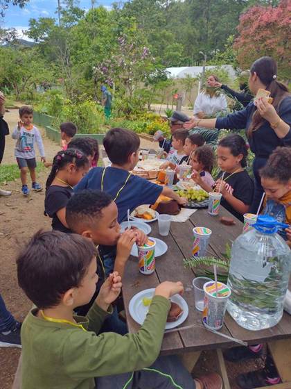 Crianças lanchando à mesa de piquenique, com suco e sanduíches, no Horto