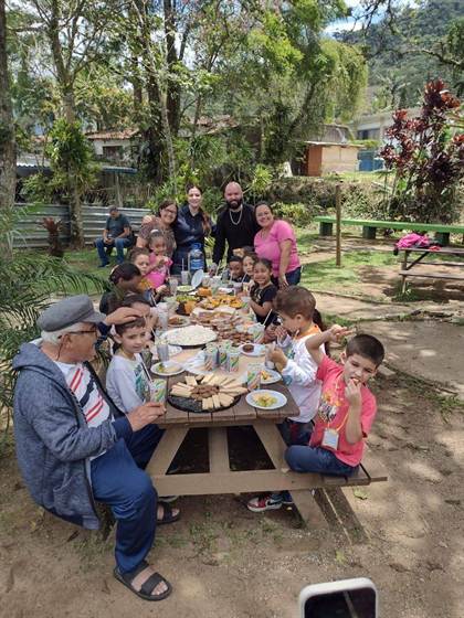 Crianças e adultos reunidos em uma mesa de piquenique com lanche variado, no Horto